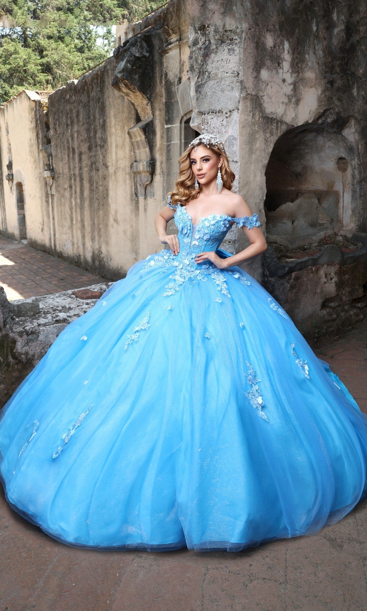 A woman poses outdoors by an old stone wall, wearing the Tiffany Blue Quinceanera Dress Q120 By Nina Canacci with off-shoulder sleeves, 3D floral details, a silver tiara, and her hand on her hip.